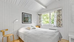 Un dormitorio con cama, una silla, una mesita de noche, un ventanal con cortinas florales y un cuadro en la pared.
