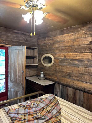 A rustic room with wooden walls, a bed with a plaid blanket, a small table with a pitcher, and a mirror on the wall.