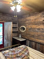 Habitación rústica con paredes de madera, una cama con manta a cuadros, una mesita con jarrón y un espejo en la pared.