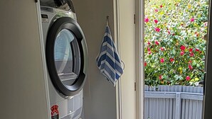 A laundry area with a washing machine and dryer, a blue laundry basket, and a door leading to a garden with a wooden fence and flowers.