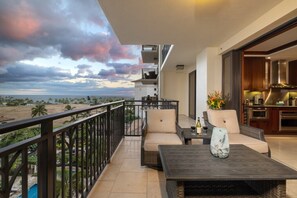 Un balcon avec vue sur la plage, un coin repas avec une table et des chaises, et une cuisine visible à travers une porte ouverte.