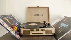 Ein alter Koffer, in dem sich ein Plattenspieler befindet, steht auf einem Tisch mit Schallplatten und einer Zeitschrift.