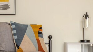 Ein Bett mit einem gemusterten Kopfkissen, ein Nachttisch mit einer Lampe und eine Wandsteckdose.