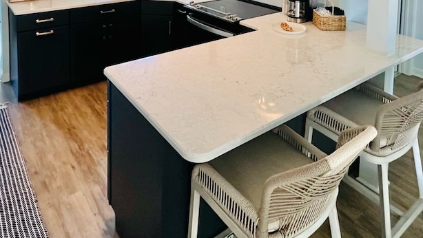 A modern kitchen with a large island, black cabinets, and a white countertop. There are two bar stools and a coffee maker on the island. - Charming 2-bedroom cottage steps away from the beach, restaurants, and downtown  (Port Austin)