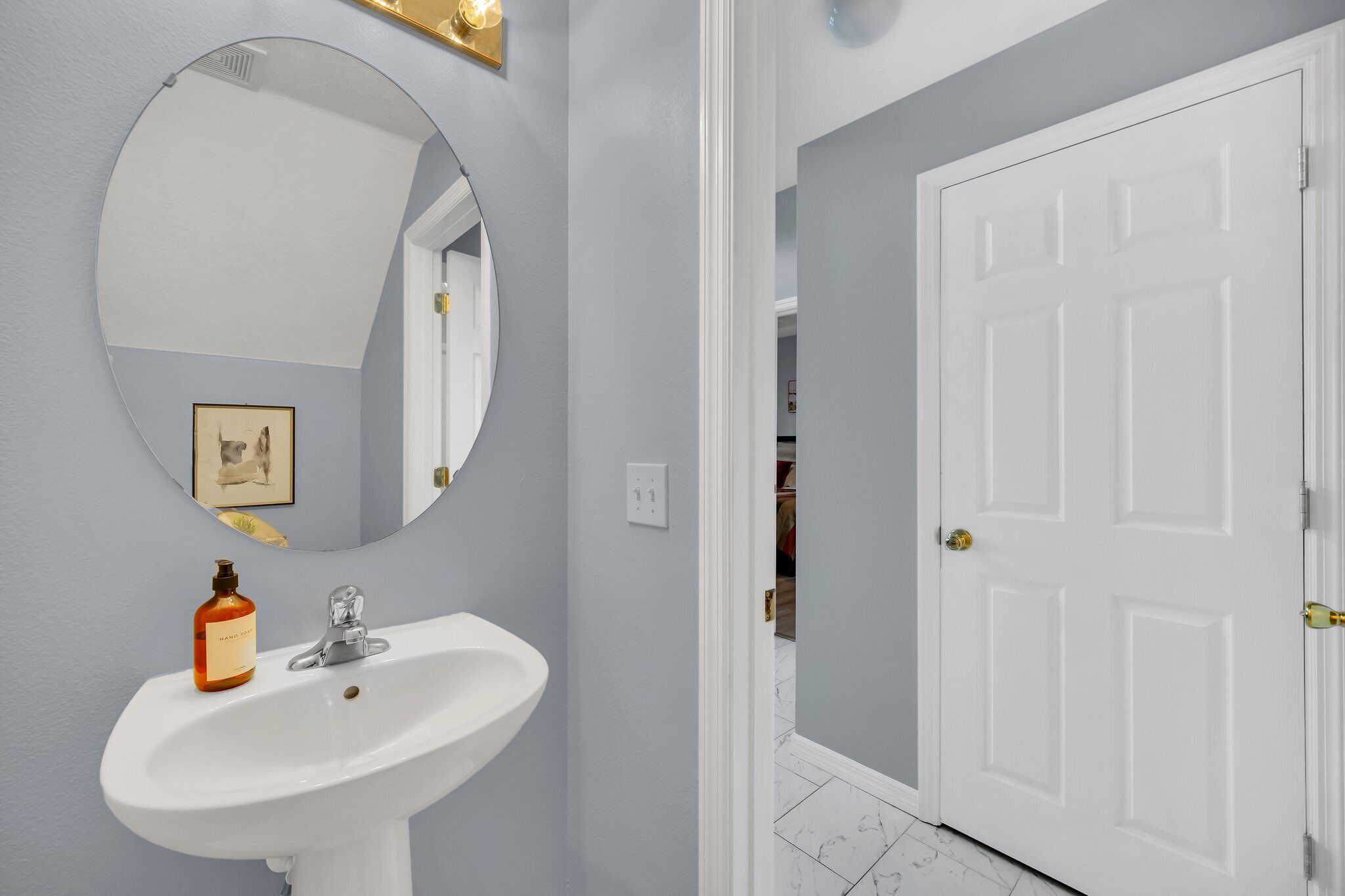 Une salle de bain avec un lavabo blanc, un miroir rond et un tableau encadré accroché au mur.