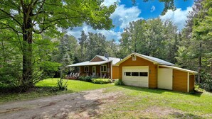 A cabin with a porch, a garage, and a dirt driveway surrounded by trees.