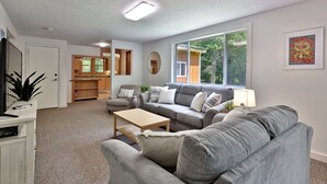 A living room with a grey sofa, a coffee table, and a TV stand. There is a kitchen area with wooden cabinets and a window.