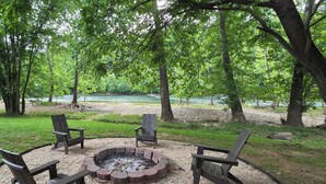 Un espace circulaire aménagé avec des sièges et un foyer, entouré de quatre chaises, situé dans une zone herbeuse avec des arbres et offrant une vue sur une rivière.