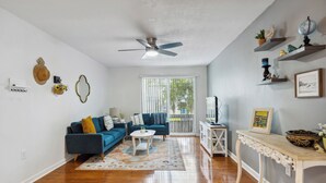 A living room with a blue sofa, a white coffee table, and a ceiling fan. - Walk to Shands - 2nd Floor Condo (Gainesville)