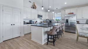 A modern kitchen with white cabinetry, a central island with bar stools, stainless steel appliances, and pendant lighting. - Coco Cabana (Hackberry)