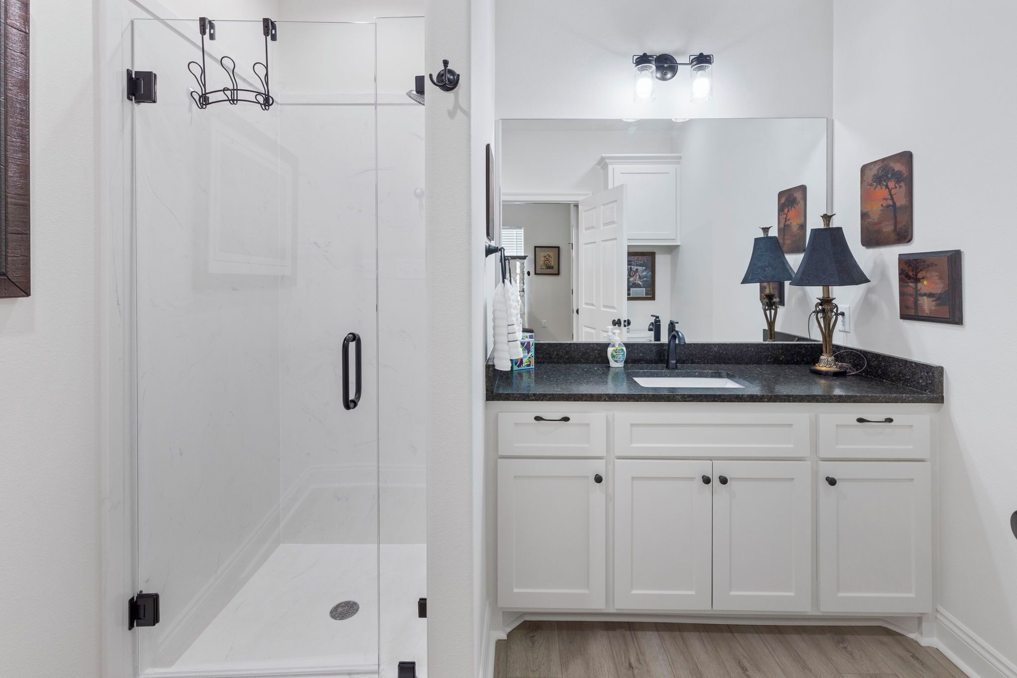 A bathroom with a glass shower door, white cabinets, a sink with a black countertop, and two wall-mounted lamps.