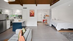 Een moderne keuken met witte kastjes, een magnetron en een inbouwoven. Er is een eethoek met een bank en een bed met een witte dekbedovertrek.
