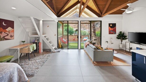 Een moderne woonkamer met een trap, een bank, een televisie en een groot raam met uitzicht op een achtertuin.