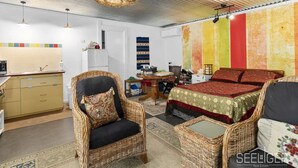 Ein Zimmer mit einem Bett, zwei Korbstühlen, einem kleinen Tisch, einer Küchenzeile und einer farbenfrohen Wand.