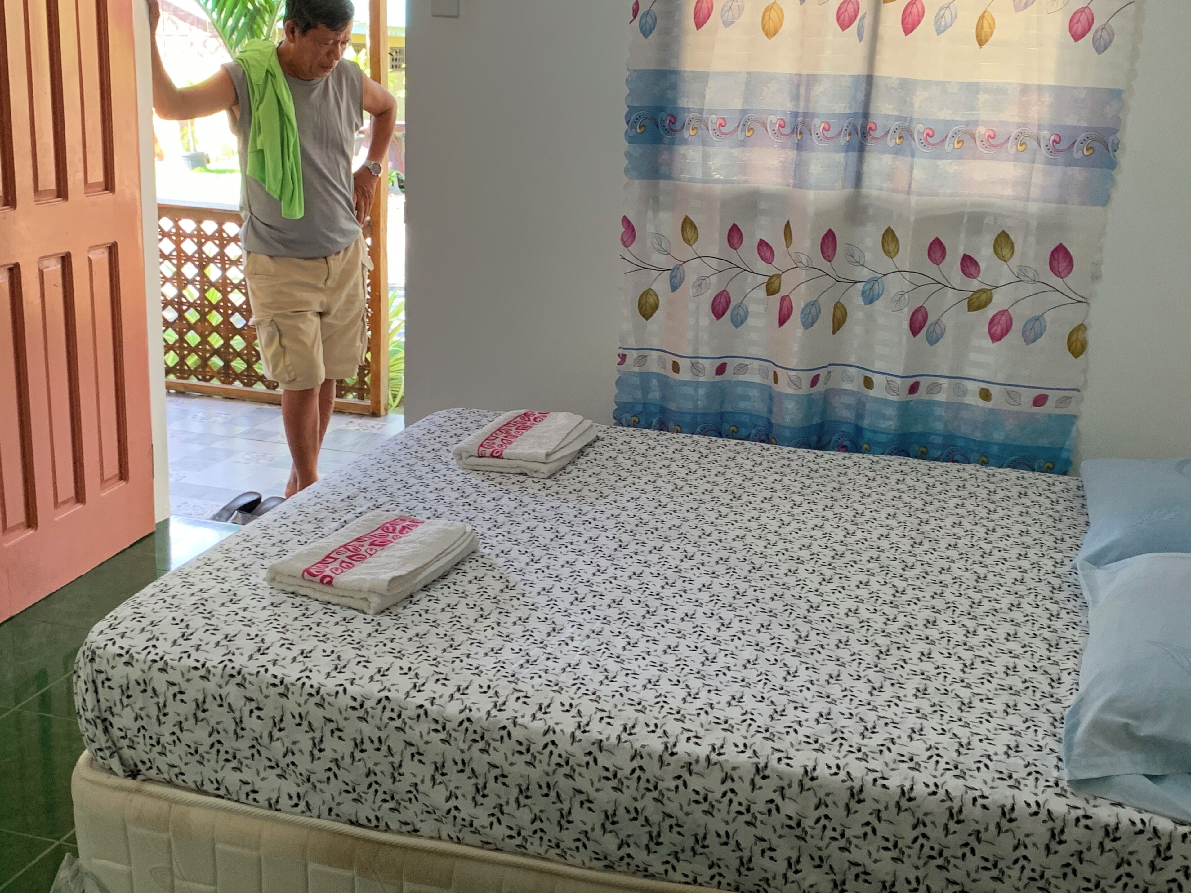 A bedroom with a bed, two towels, a green towel draped over a door, and a colorful curtain.