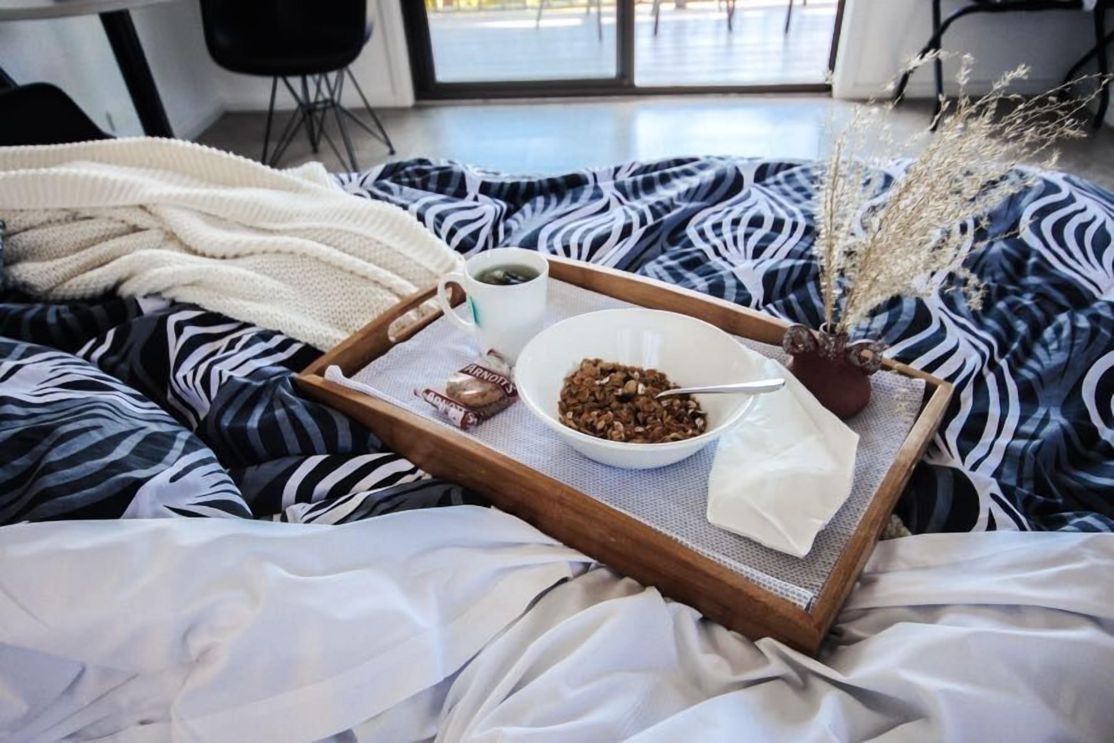 Un plateau avec un bol de céréales, une tasse de café et un petit paquet, posé sur un lit recouvert d’une couverture à rayures zébrées.