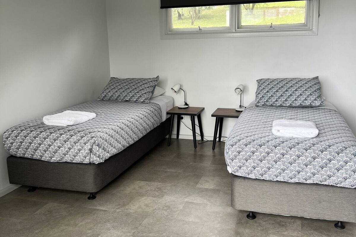Ein Schlafzimmer mit zwei Betten, jeweils mit gemusterten Bettdecken und Kissen, einem kleinen Tisch und einem Fenster mit Verdunkelungsvorhang.