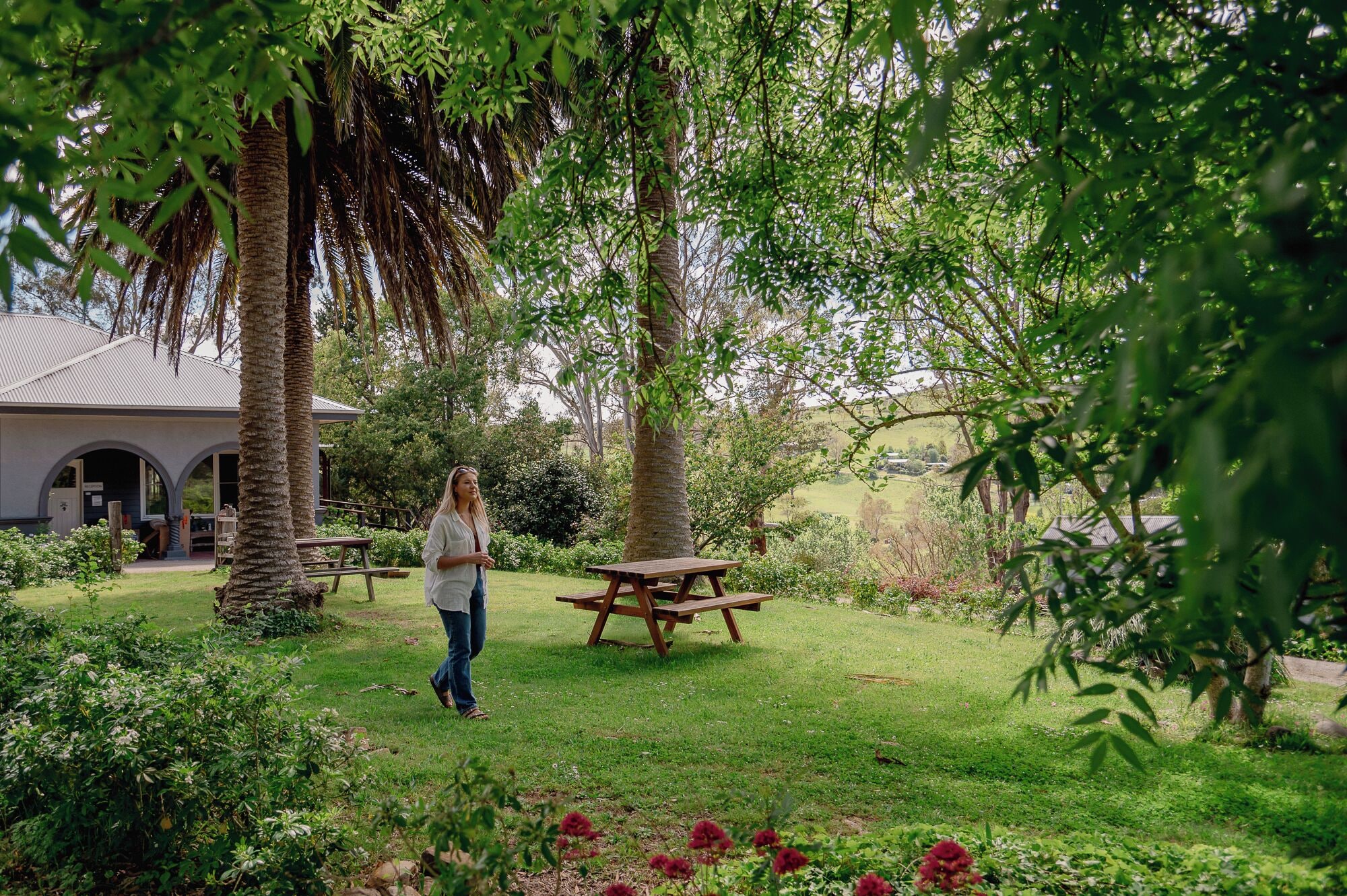 A woman walking in a garden with picnic tables and a building in the background.