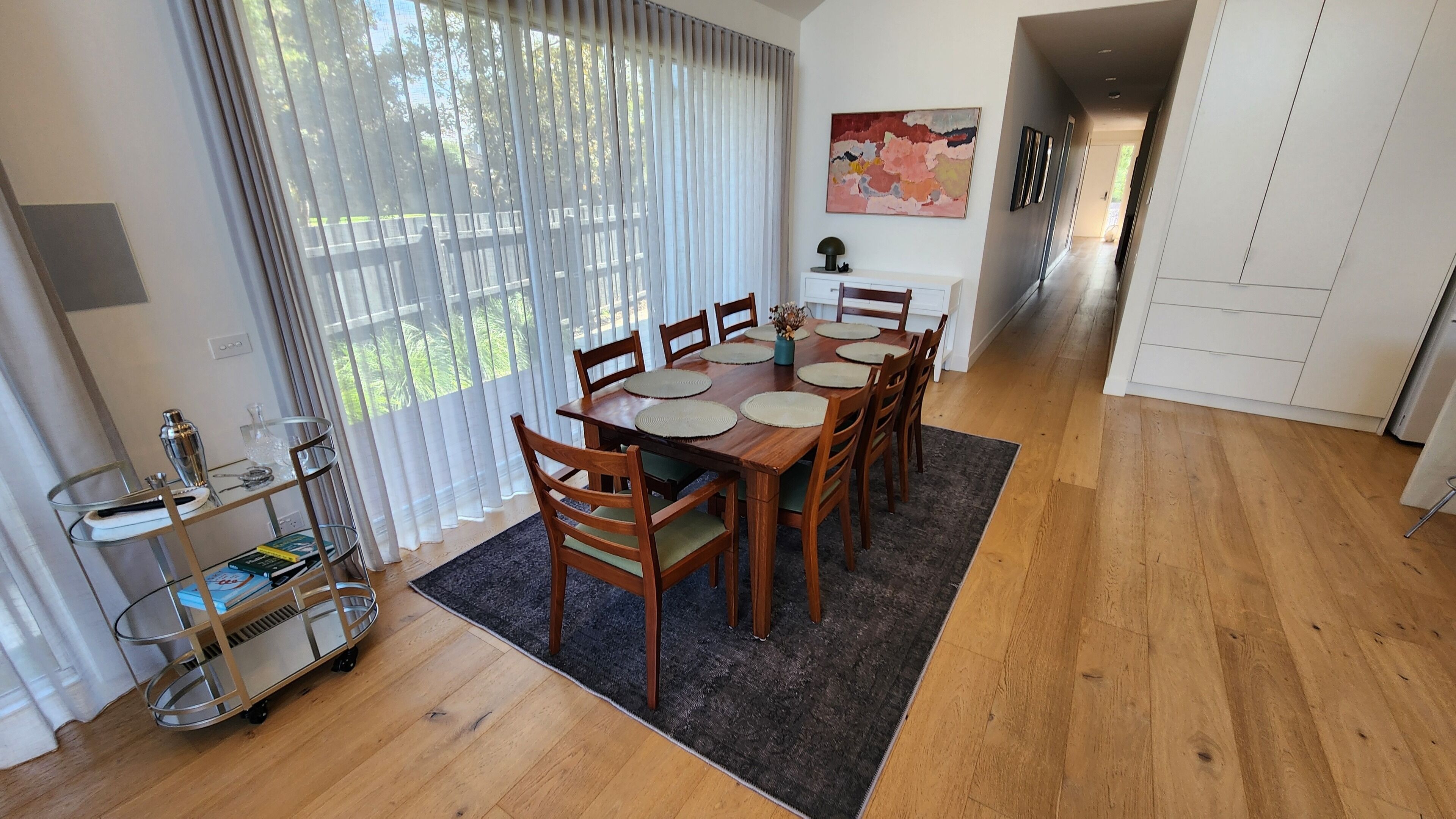 A dining area with a wooden table, chairs, and a rug. There is a window with curtains, a metal cart with items, and a painting on the wall.