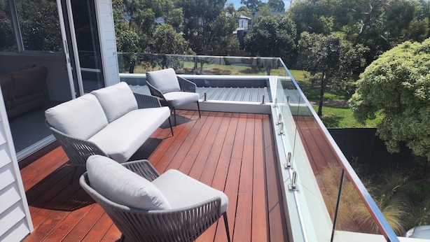 A wooden deck with outdoor furniture, including a sofa and chairs, overlooking a garden with trees and a house in the background. - Luxury Coastal Townhouse Mornington (Mornington)