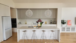 A modern kitchen with a central island, bar stools, a built-in oven, and a refrigerator. - Luxury Coastal Townhouse Mornington (Mornington)