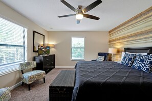 Une chambre à coucher comprenant un lit, un ventilateur de plafond, une commode, une chaise et une fenêtre avec des stores.