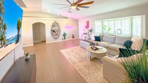 A spacious living room with a ceiling fan, a large window with blinds, a sofa, a coffee table, and a painting on the wall. - Hav-A-Time! Pet Friendly Pool House w/ Game Room! (Lake Havasu City)