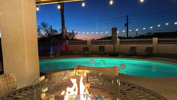 A poolside area with a glass table and fire pit, surrounded by lounge chairs and string lights. - Hav-A-Time! Pet Friendly Pool House w/ Game Room! (Lake Havasu City)