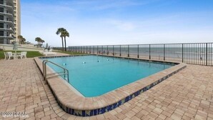 Una piscina rectangular con borde de azulejos, rodeada por un patio de ladrillo y una valla metálica.