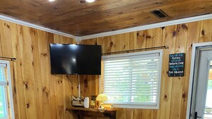 Une pièce chaleureuse aux murs en bois, avec un téléviseur à écran plat fixé au-dessus d’une étagère, deux canapés marron, un tapis à motifs et une fenêtre avec des stores.