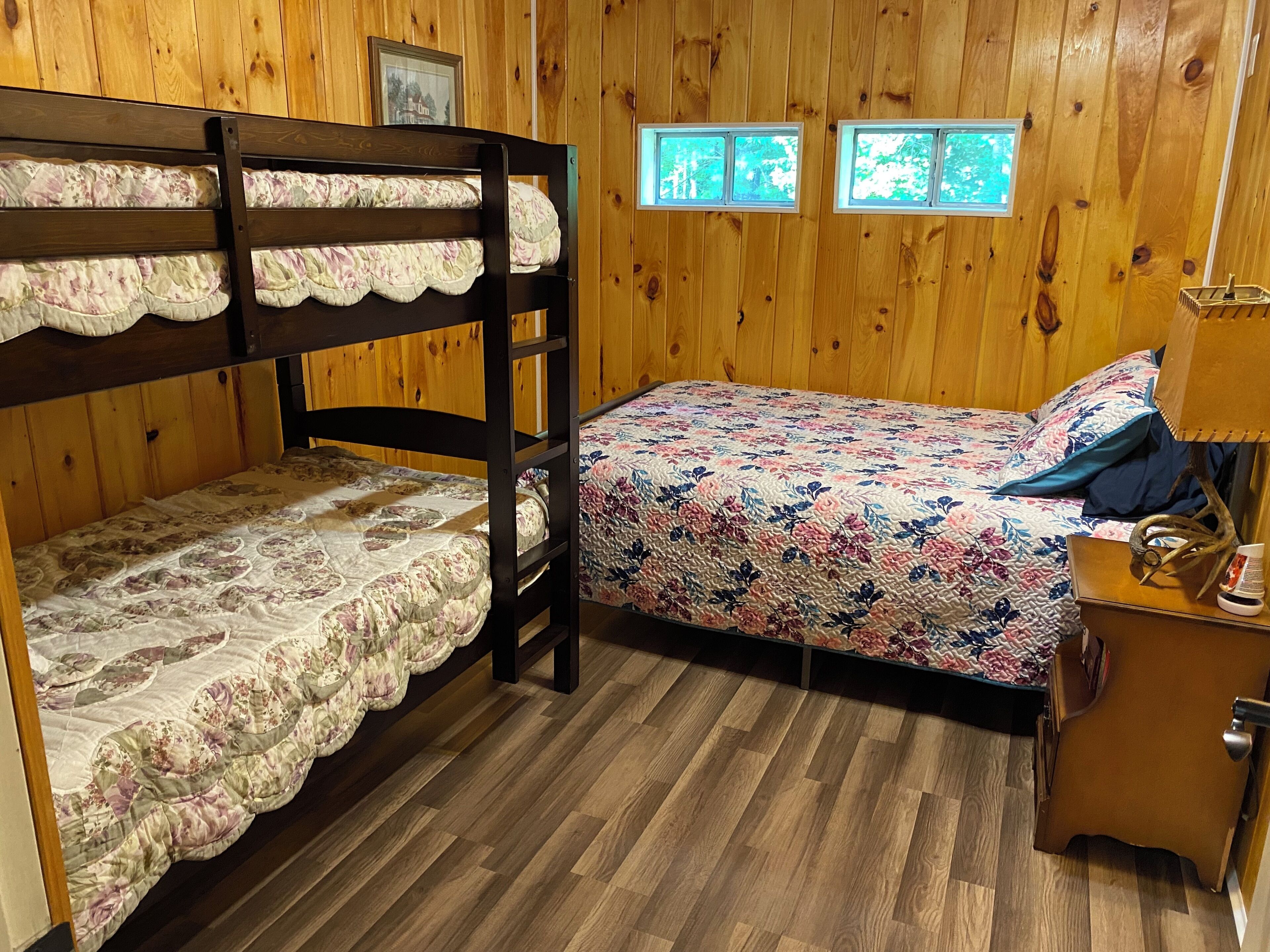 Un lit superposé avec une literie à motifs floraux, un lit simple avec une courtepointe à motifs floraux, des murs en bois et une table de chevet avec une lampe.