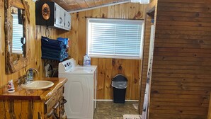 Une salle de bain rustique aux murs en bois et au sol carrelé, comprenant un lavabo, un lave-linge et une fenêtre avec des stores.