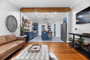 Un salon moderne avec un canapé en cuir marron, un pouf à motifs et une table basse en bois. On aperçoit, au fond, une cuisine avec des meubles bleus et un îlot central.