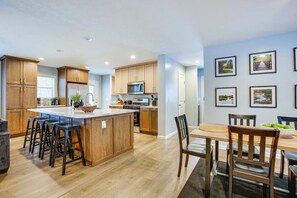 Une cuisine moderne avec des meubles en bois, un îlot central doté de tabourets, un coin repas avec une table et des chaises, et des tableaux encadrés accrochés au mur.