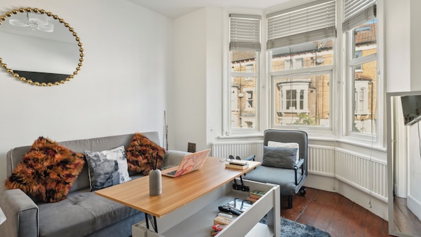A modern living room with a grey sofa, a wooden coffee table, and a view of buildings through a bay window. - Beautiful Abode in Brixton (London)