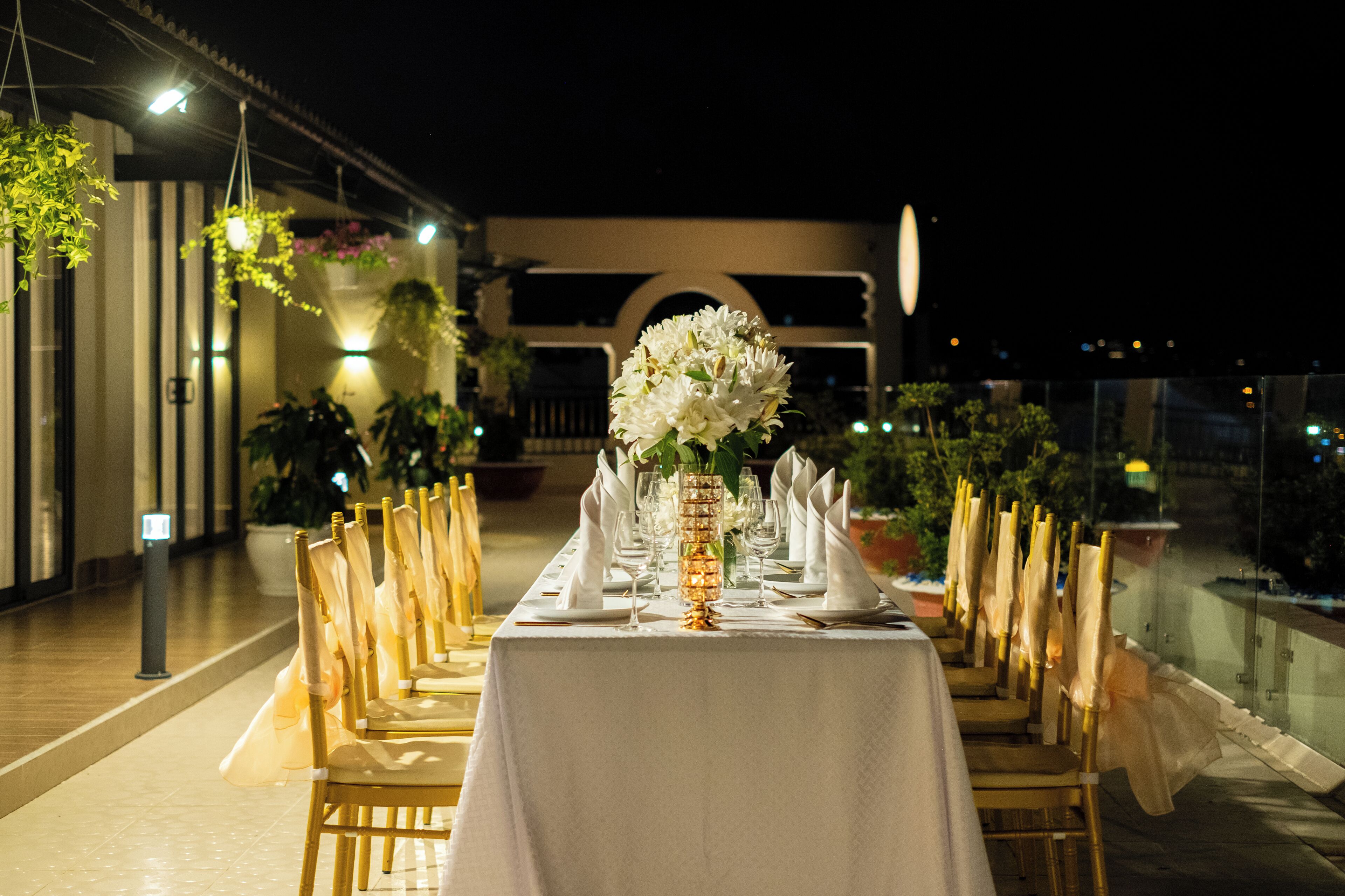 Une table de banquet dressée pour un événement formel, avec des nappes blanches, des chaises dorées et un centre de table composé de fleurs blanches.