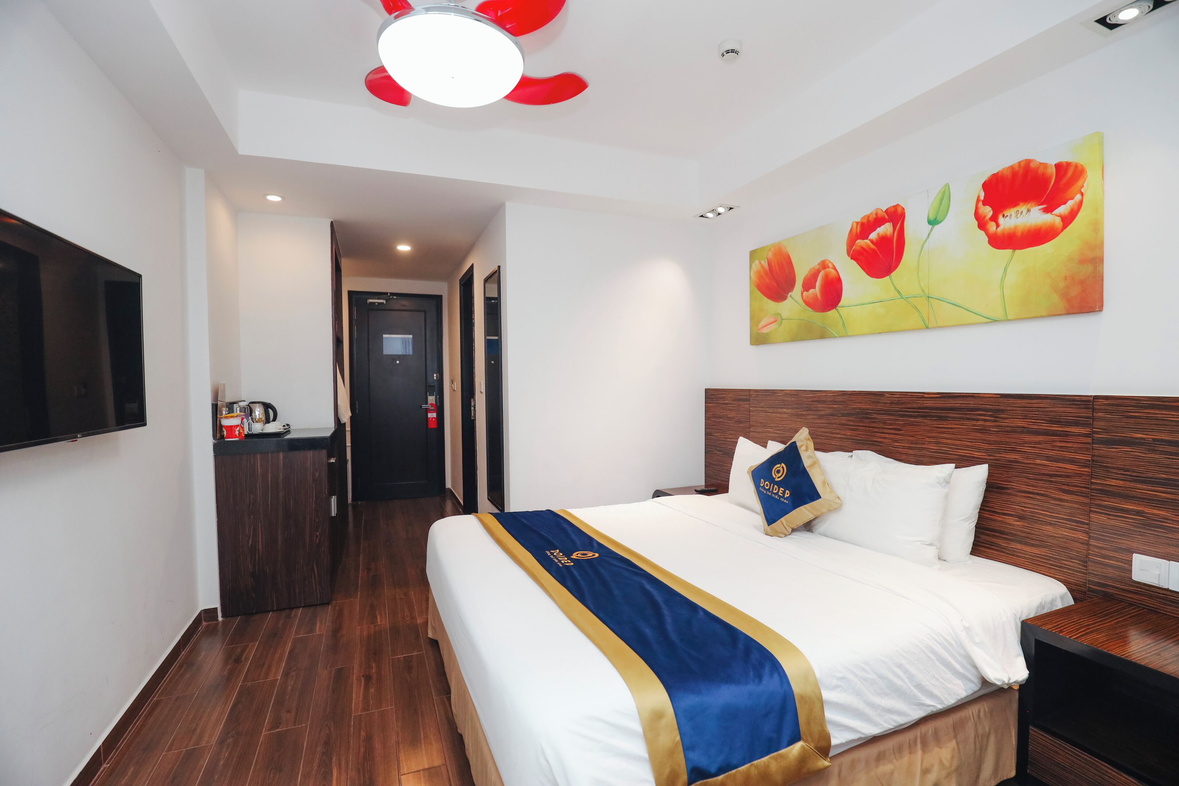 A hotel room with a large bed, a flat-screen TV, a painting of red flowers, and a wooden headboard.