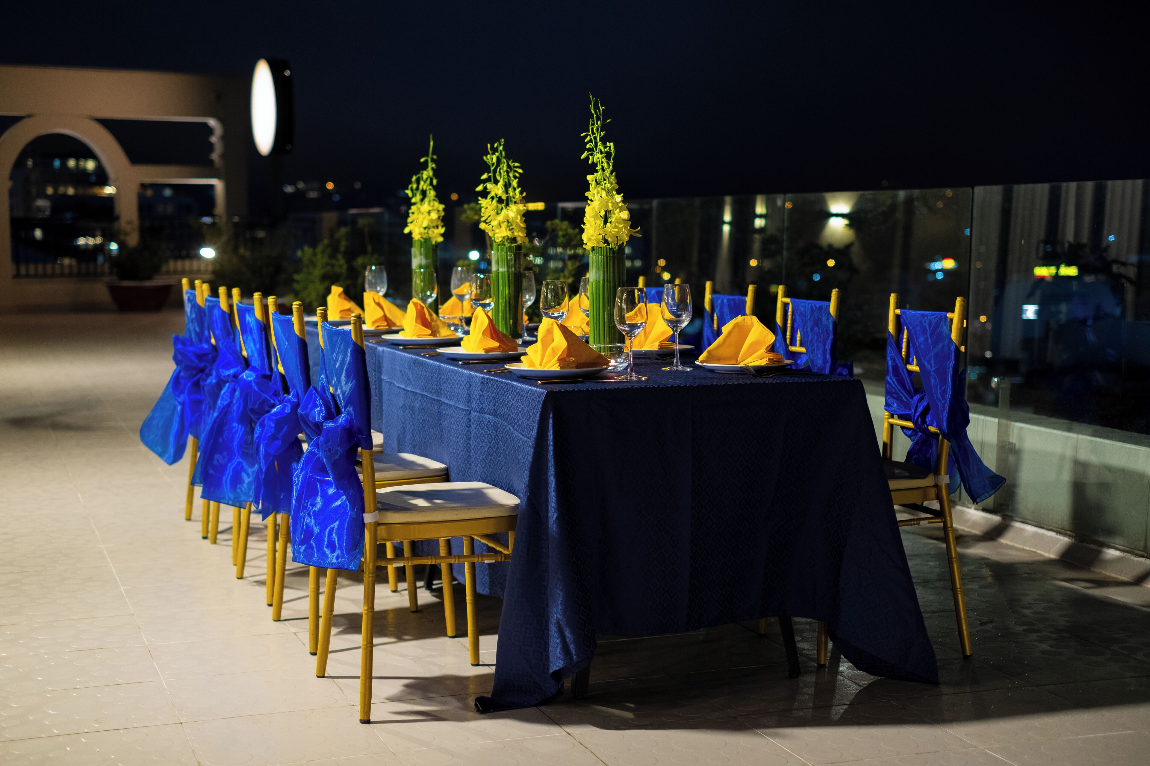 A table set for a meal with blue tablecloths and yellow napkins, chairs with blue sashes, and yellow flowers in vases.