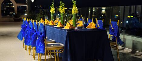 A table set for a meal with blue tablecloths and yellow napkins, chairs with blue sashes, and yellow flowers in vases.