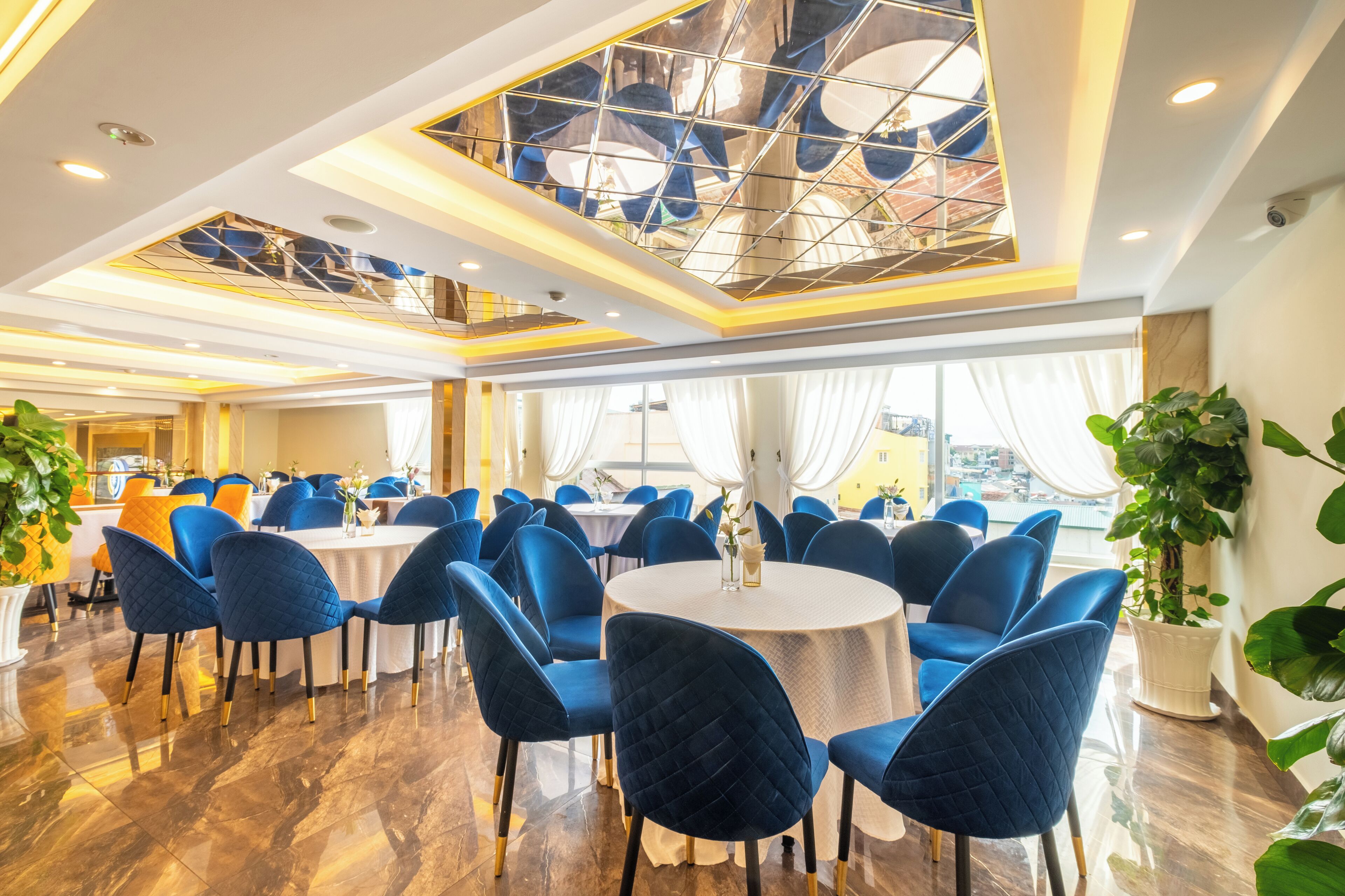 A spacious dining area with round tables and blue upholstered chairs, a high ceiling with geometric patterns, and large windows allowing natural light.