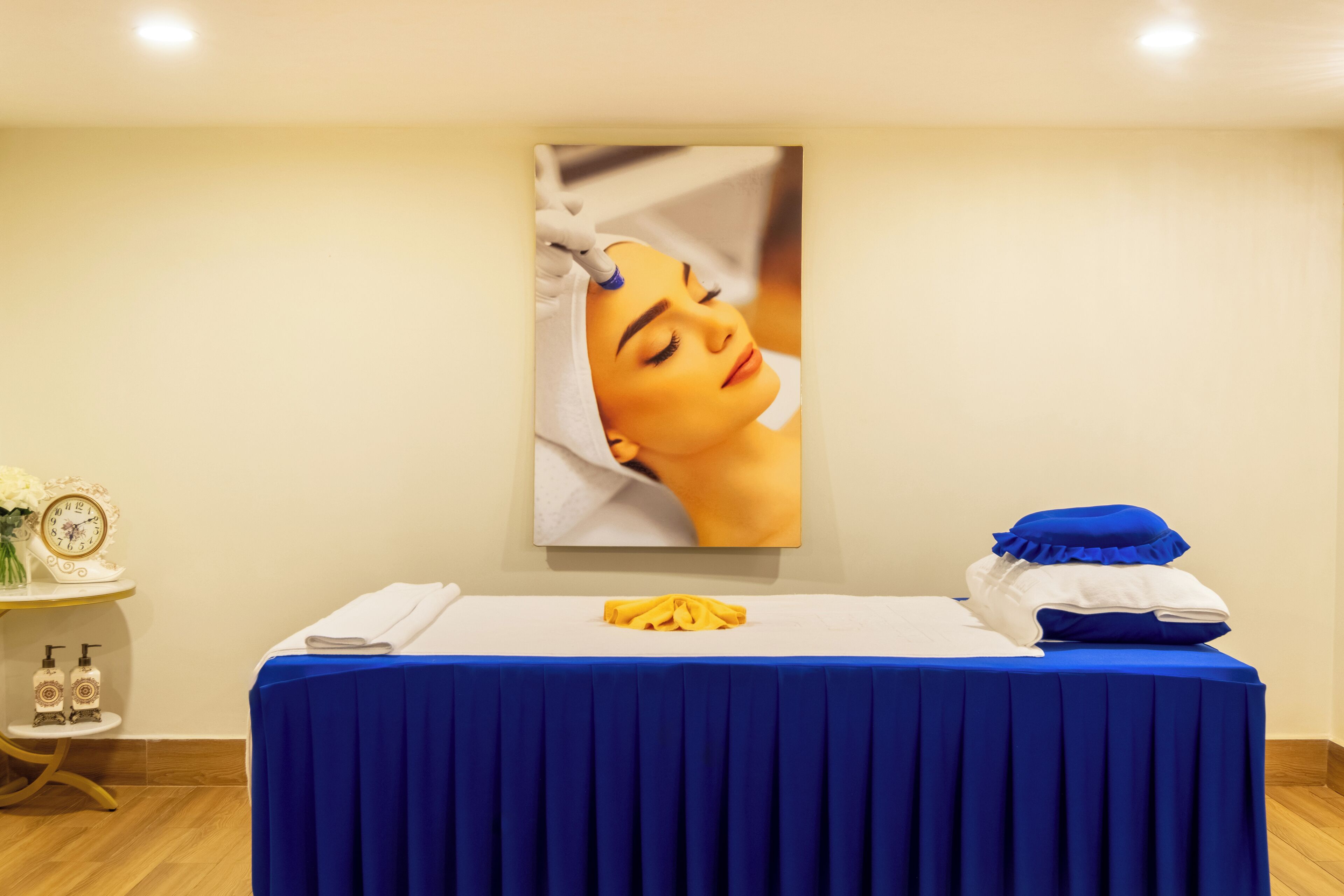 A room with a blue-covered massage table, a clock, and a vase of flowers on a side table.