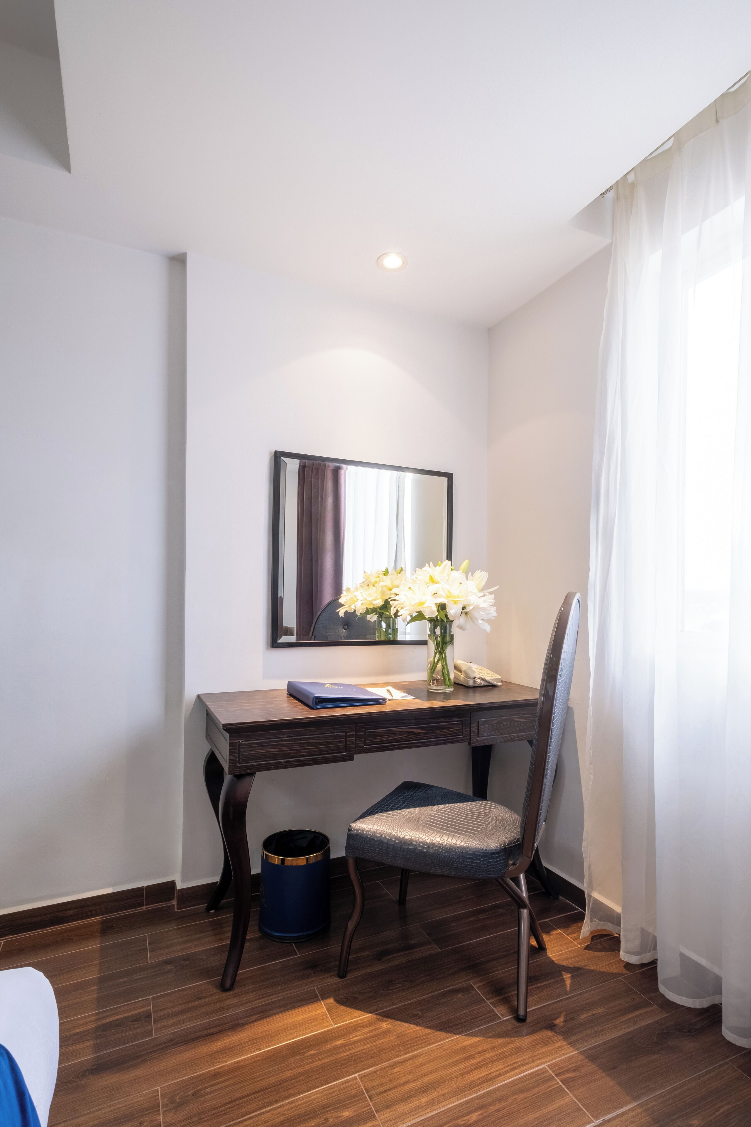 A hotel room with a wooden desk, a chair, a vase with flowers, a mirror, and a window with sheer curtains.