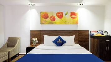A hotel room with a bed, a chair, a nightstand, and a painting of flowers on the wall.