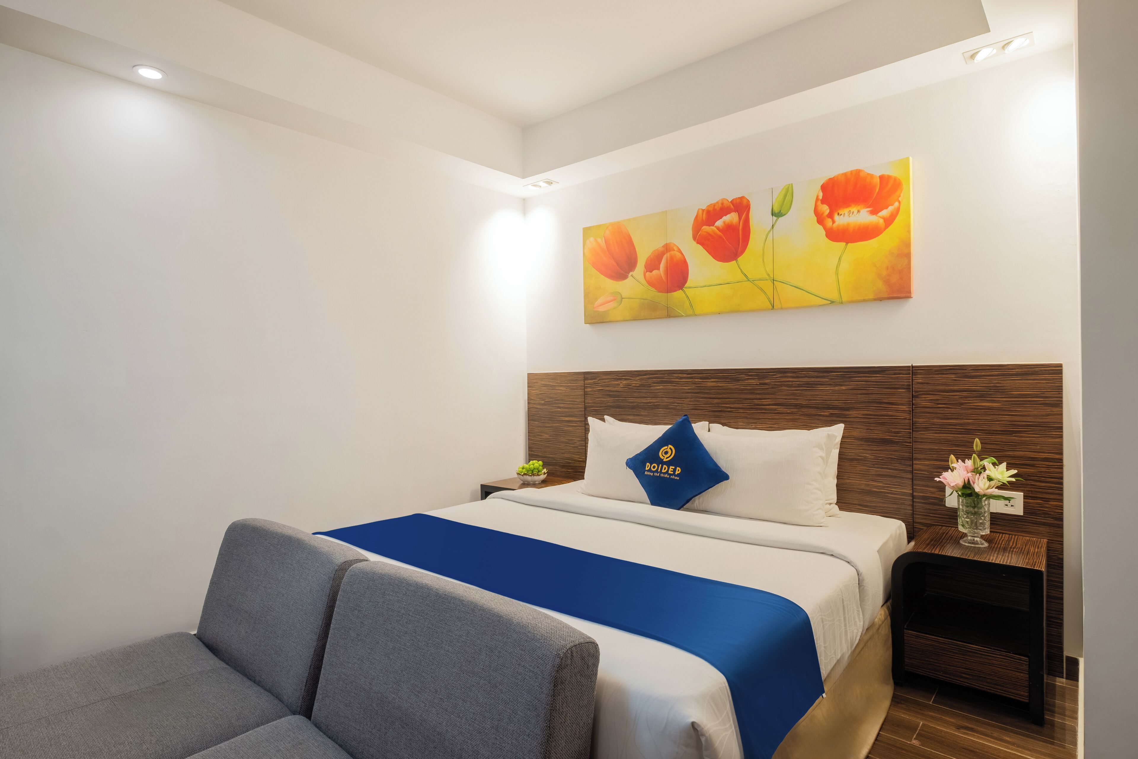 A hotel room with a bed, a blue bedspread, a wooden headboard, a nightstand with a vase of flowers, and a painting of flowers on the wall.