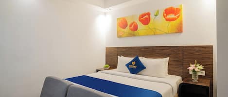 Habitación de hotel con cama, cubrecama azul, cabecera de madera, mesita de noche con un florero y un cuadro de flores en la pared.