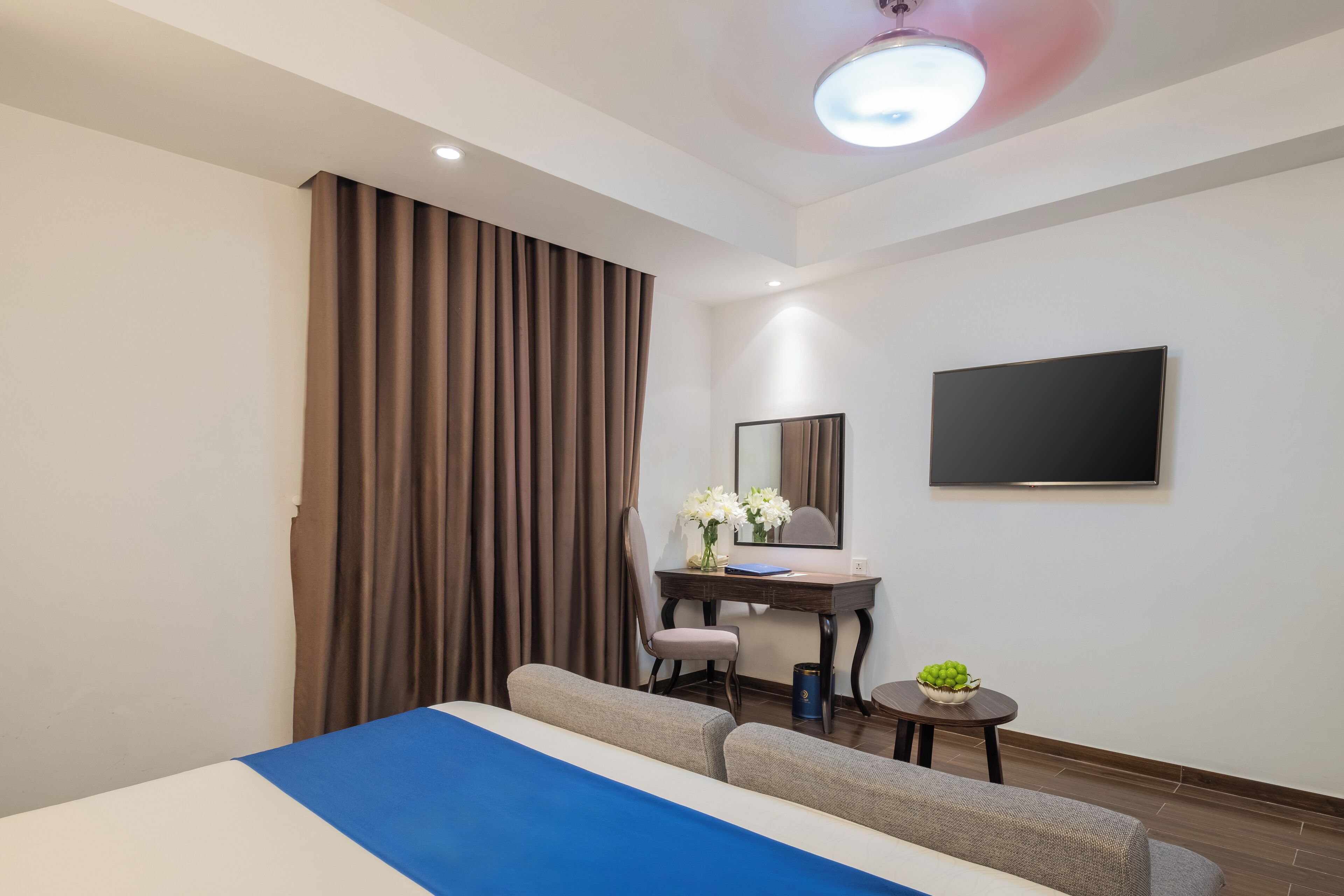 Een hotelkamer met een bed, een bureau, een stoel, een bijzettafeltje met een plant en een flatscreen televisie aan de muur.
