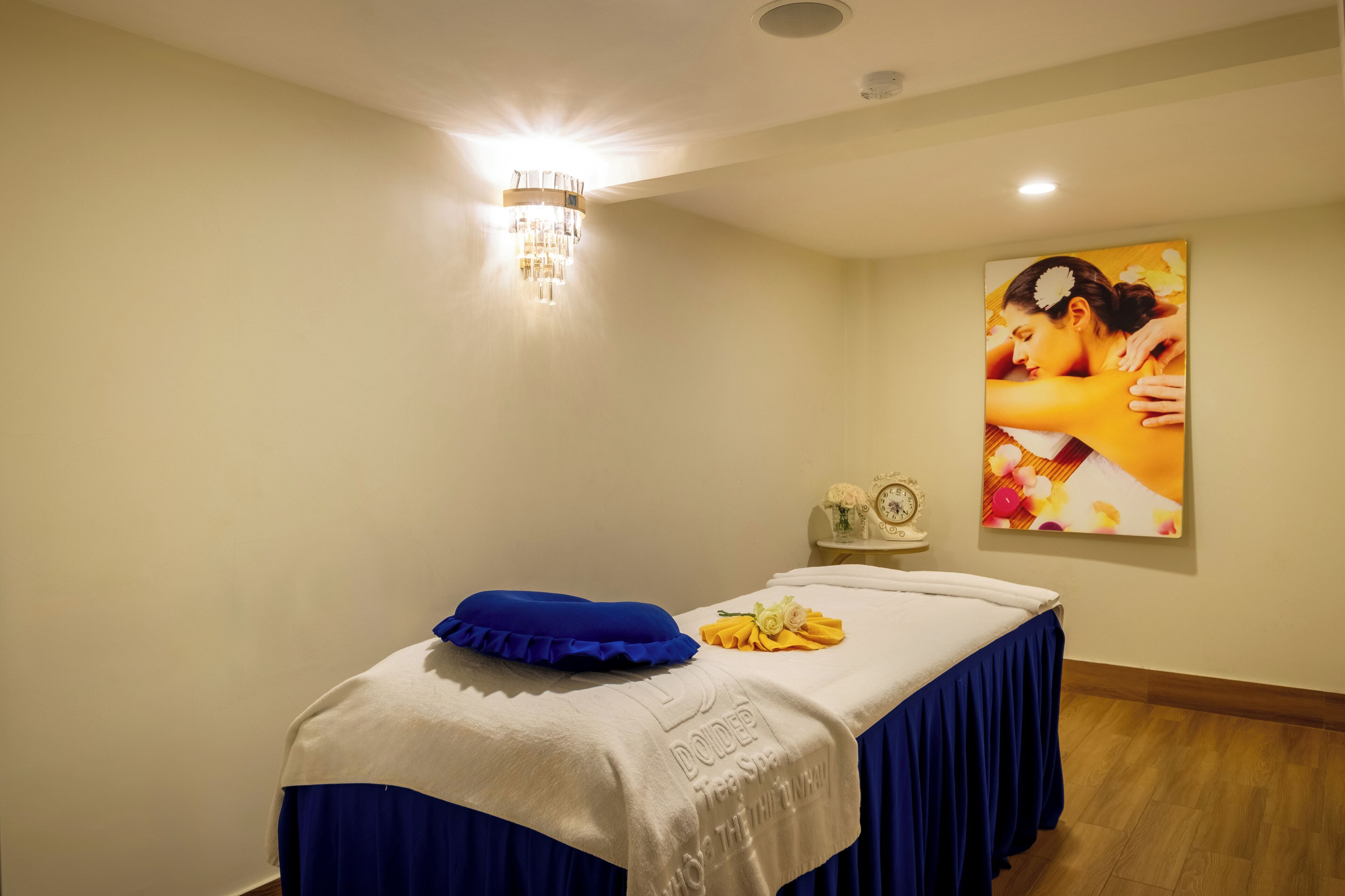 A massage room with a massage table, a blue pillow, a tray with a towel and flowers, a wall clock, and a poster.