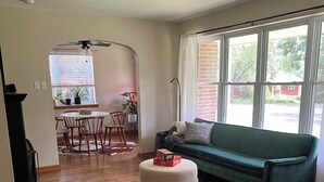 Ein Wohnzimmer mit einem grünen Sofa, einem grünen Sessel, einem Esstisch mit Stühlen und einem großen Fenster mit Jalousien.
