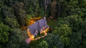 Une vue aérienne d’une maison à deux étages, entourée d’une forêt dense, avec une allée bien éclairée et un éclairage extérieur.