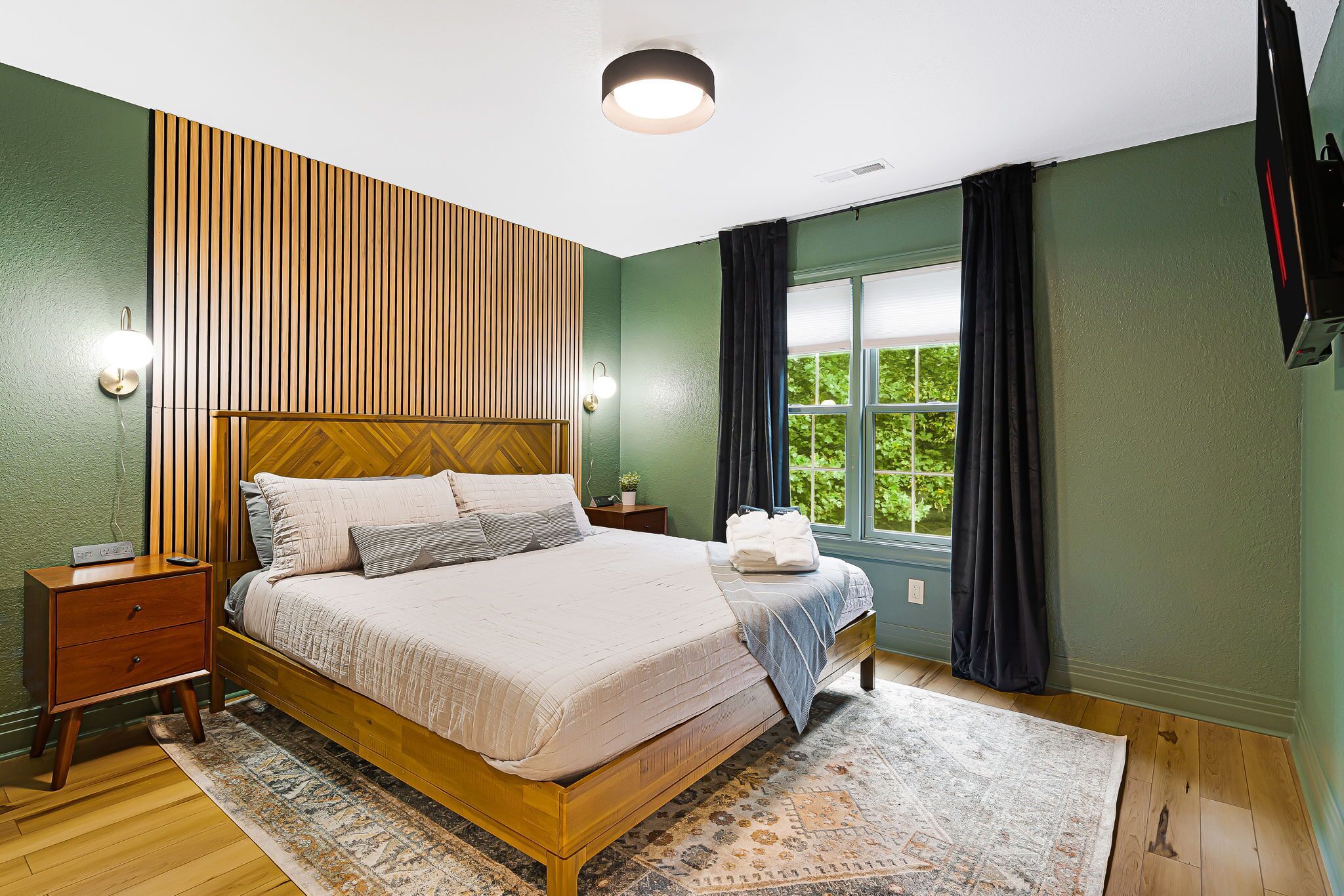 Une chambre avec un lit en bois, une table de chevet, une lampe fixée au mur, une fenêtre avec des rideaux et une vue sur la verdure à l’extérieur.
